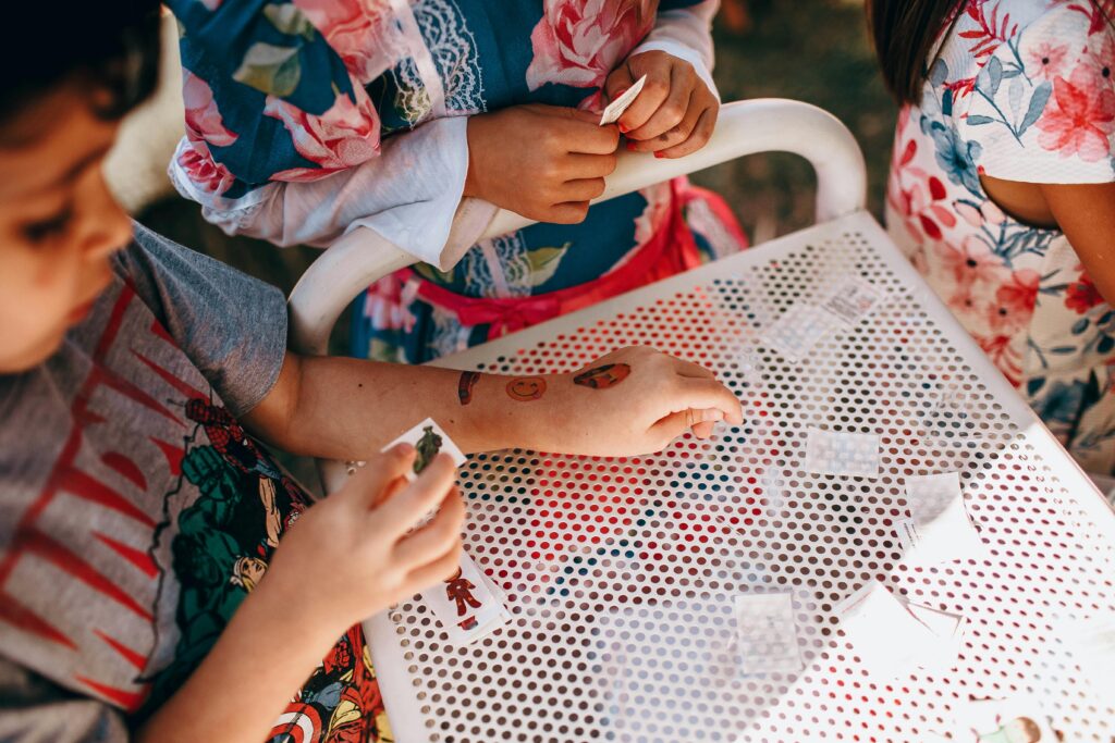 From above of anonymous kids in casual clothes standing near white table and playing with small cards in sunny day